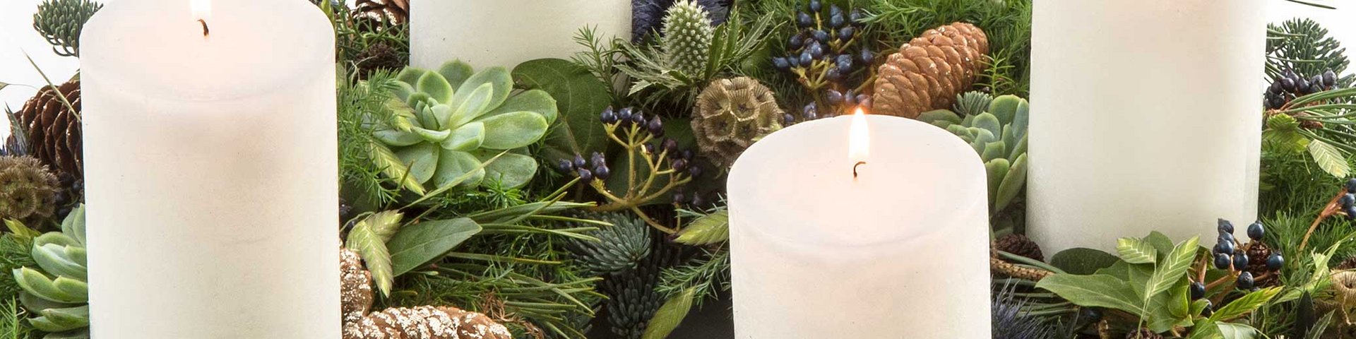 En dekorativ adventskrans med fire hvide stearinlys, der er omgivet af grønne planter, kogler og bær. Stearinlysene er tændt og skaber en hyggelig atmosfære. Kransen er fyldig og prydet med naturmaterialer.