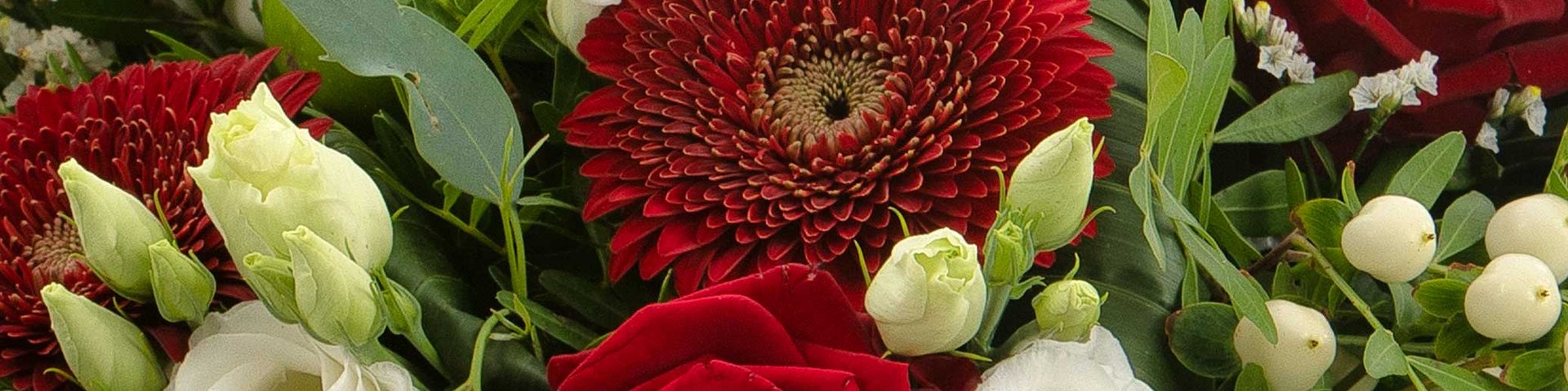 Et farverigt blomsterbuket med røde roser, hvide lysser og en dyb rød gerbera i midten. Grønne blade og små bær er også til stede, hvilket skaber en frodig og livlig sammensætning.